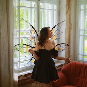 May include: A woman wearing a black off-the-shoulder dress and fairy wings with yellow and orange accents. The wings are attached to a black frame. The background includes a window, curtains, and a red armchair.