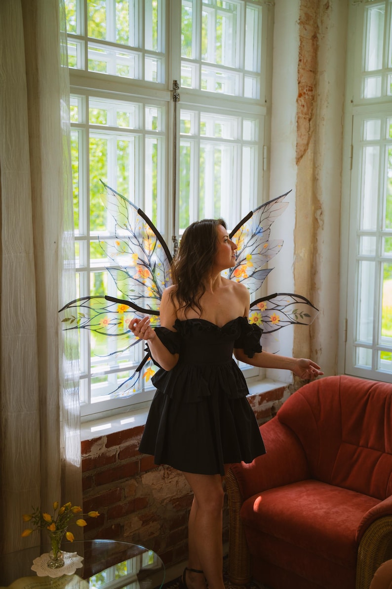 May include: A woman wearing a black off-the-shoulder dress and fairy wings with yellow and orange accents. The wings are attached to a black frame. The background includes a window, curtains, and a red armchair.