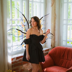 May include: A woman wearing a black off-the-shoulder dress and fairy wings stands near a window. The wings are translucent with black outlines and colorful accents. A red armchair is in the background.