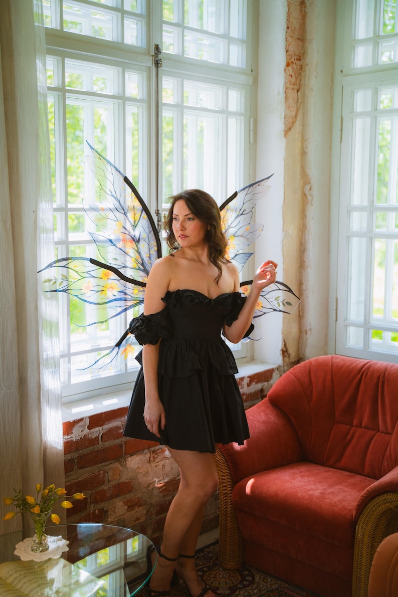 May include: A woman wearing a black off-the-shoulder dress and fairy wings stands near a window. The wings are translucent with black outlines and colorful accents. A red armchair is in the background.