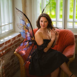 May include: A woman wearing a black dress and fairy wings with a colorful design, seated in a red armchair. The wings have a translucent appearance with blue, yellow, and orange accents. The background includes windows and a brick wall.