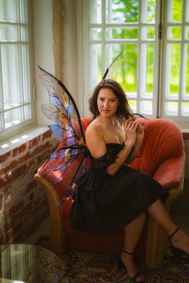 May include: A woman wearing a black dress and fairy wings with a colorful design, seated in a red armchair. The wings have a translucent appearance with blue, yellow, and orange accents. The background includes windows and a brick wall.