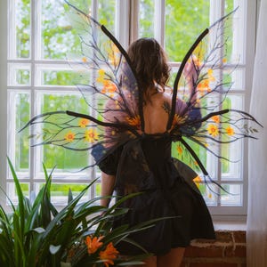 May include: A person wearing a black dress and transparent fairy wings with yellow and orange floral accents. The wings have black branch-like structures. The person is standing in front of a window with a view of greenery.