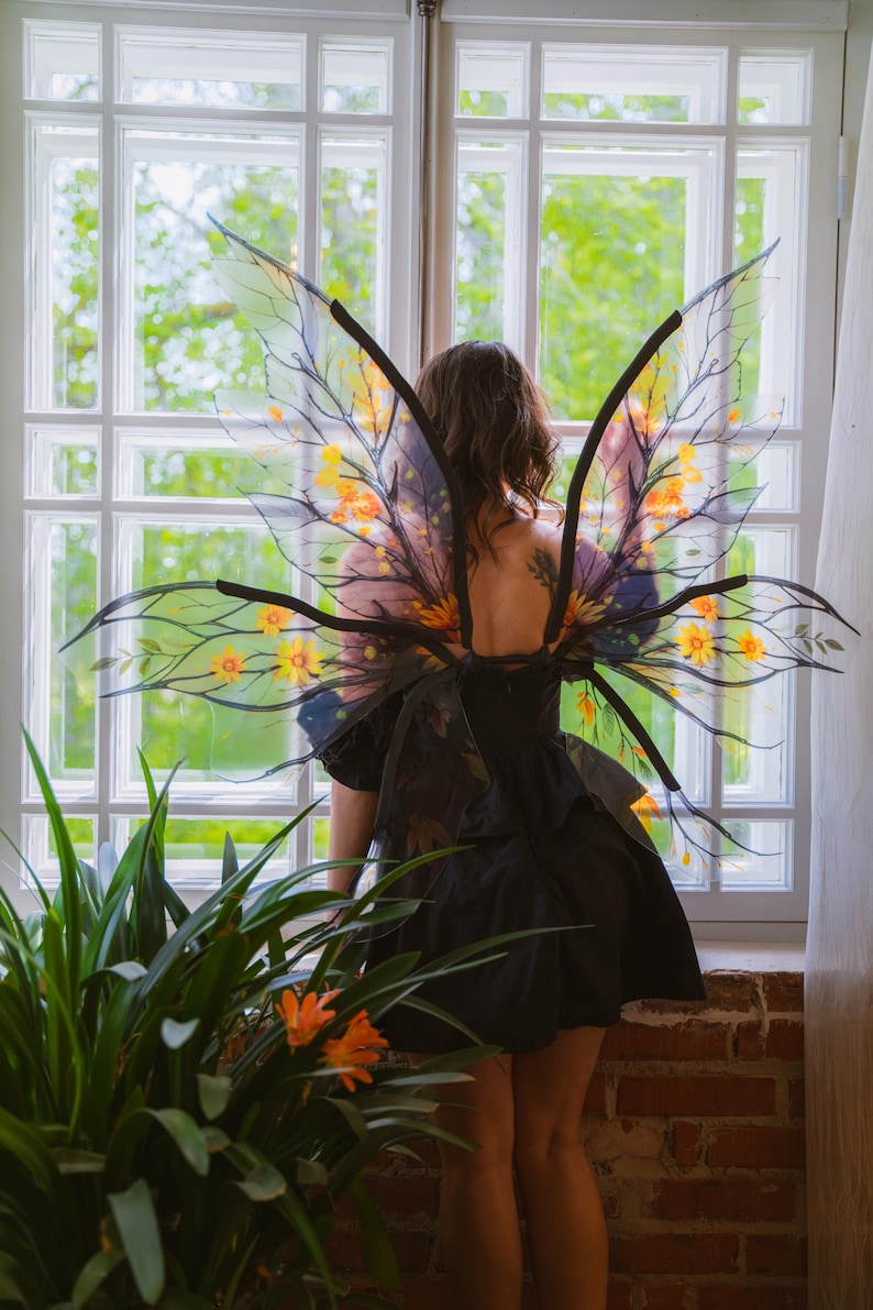 May include: A person wearing a black dress and transparent fairy wings with yellow and orange floral accents. The wings have black branch-like structures. The person is standing in front of a window with a view of greenery.