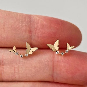 以下が含まれることがあります： 小さな透明な石が付いた金の蝶のイヤリングのペア。各イヤリングには、蝶が2匹と石が3つ付いています。