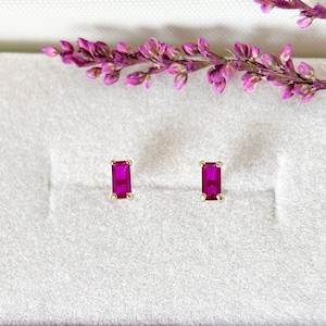 May include: A pair of gold-toned stud earrings, each featuring a rectangular, vibrant magenta gemstone. The earrings are set against a soft, light gray background, with a sprig of purple flowers in the upper right corner.