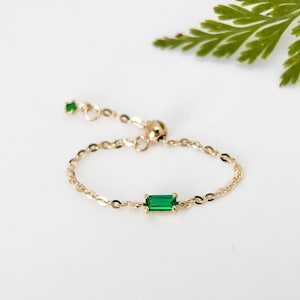 Puede incluir: Un delicado anillo de cadena de oro con una piedra preciosa de esmeralda verde. El anillo es ajustable y tiene un pequeño acento de esmeralda verde en el lateral.