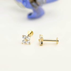 Puede incluir: Un pendiente de flor de circonita cúbica de cuatro puntas, engastado en garras, de color dorado. El pendiente se muestra sobre un fondo blanco.