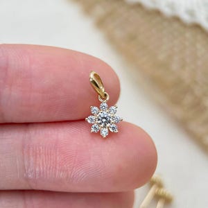 May include: A small gold pendant featuring a central round brilliant-cut diamond surrounded by eight smaller round diamonds arranged in a floral starburst pattern, complete with a simple gold bail at the top for attachment to a necklace chain.