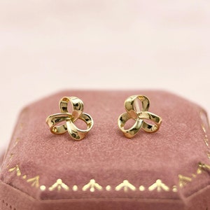 Może przedstawiać: Para złotych kolczyków sztyftów, każdy z trój pętlowym wzorem. Kolczyki są prezentowane na różowym aksamitnym pudełku na biżuterię ze złotymi wykończeniami, na tle miękkiego różowego tła. Kolczyki mają wypolerowaną, odblaskową powierzchnię.