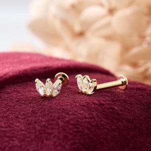 May include: Two gold-colored, stud earrings with a crown-like design, each featuring three clear, marquise-cut gemstones. The earrings are displayed on a deep red, textured fabric, with a soft, blurred background.