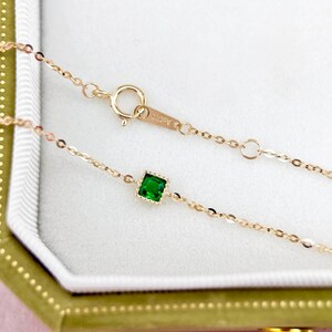 May include: A delicate gold chain necklace with a small square green gemstone pendant. The chain is a delicate, fine link style. The pendant is set in a gold frame.