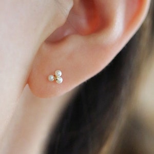 May include: A close-up of a gold stud earring featuring three tiny, white pearls in a cluster design. The earring is set against the backdrop of a person's ear, highlighting the jewellery's delicate nature.