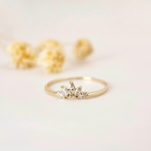 Puede incluir: Un delicado anillo de oro con un dise&ntilde;o de corona con cinco piedras preciosas claras con corte marquesa. El anillo se presenta sobre un fondo blanco suave, mostrando su estilo elegante y minimalista.