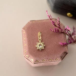 May include: A gold pendant featuring a central clear stone surrounded by a halo of smaller clear stones in a starburst arrangement. The pendant has a small bail at the top and is resting on a dusty rose velvet octagonal jewelry box.