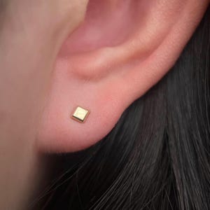 May include: A close-up of a person's ear with a small, square-shaped gold earring.
