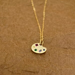 Op de afbeelding: Een gouden ketting met een klein, kleurrijk palet-hanger. Het palet is versierd met rode, gele, groene, blauwe en paarse edelstenen.
