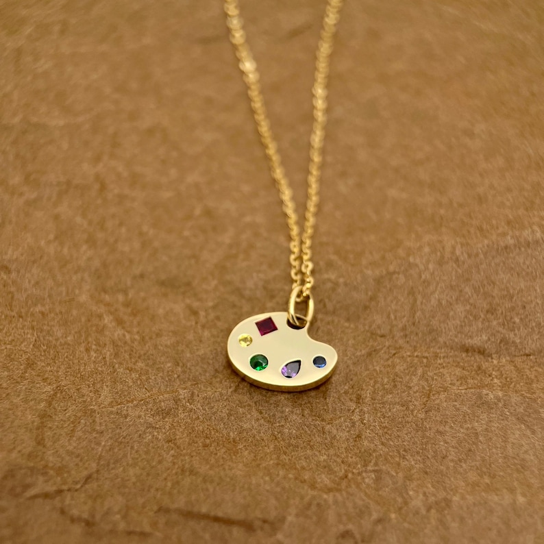 Op de afbeelding: Een gouden ketting met een klein, kleurrijk palet-hanger. Het palet is versierd met rode, gele, groene, blauwe en paarse edelstenen.