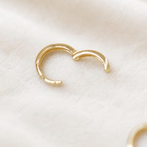 May include: A gold-colored hoop earring, partially open, against a soft, white background. The earring has a simple, rounded design. The image is well-lit, highlighting the earring's smooth surface and the clasp mechanism.