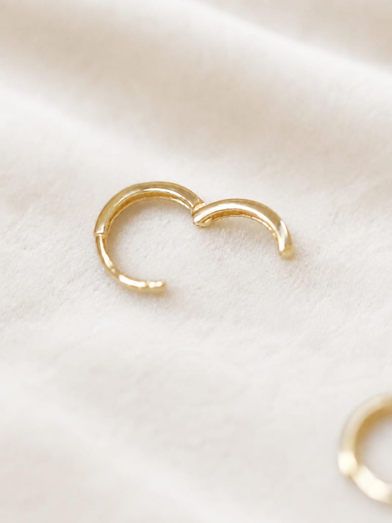 May include: A gold-colored hoop earring, partially open, against a soft, white background. The earring has a simple, rounded design. The image is well-lit, highlighting the earring's smooth surface and the clasp mechanism.
