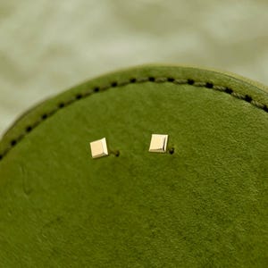 May include: A pair of small, square-shaped gold earrings. The earrings are on a green background.