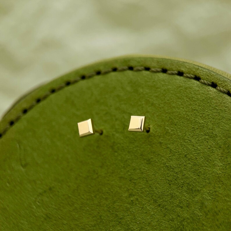 May include: A pair of small, square-shaped gold earrings. The earrings are on a green background.