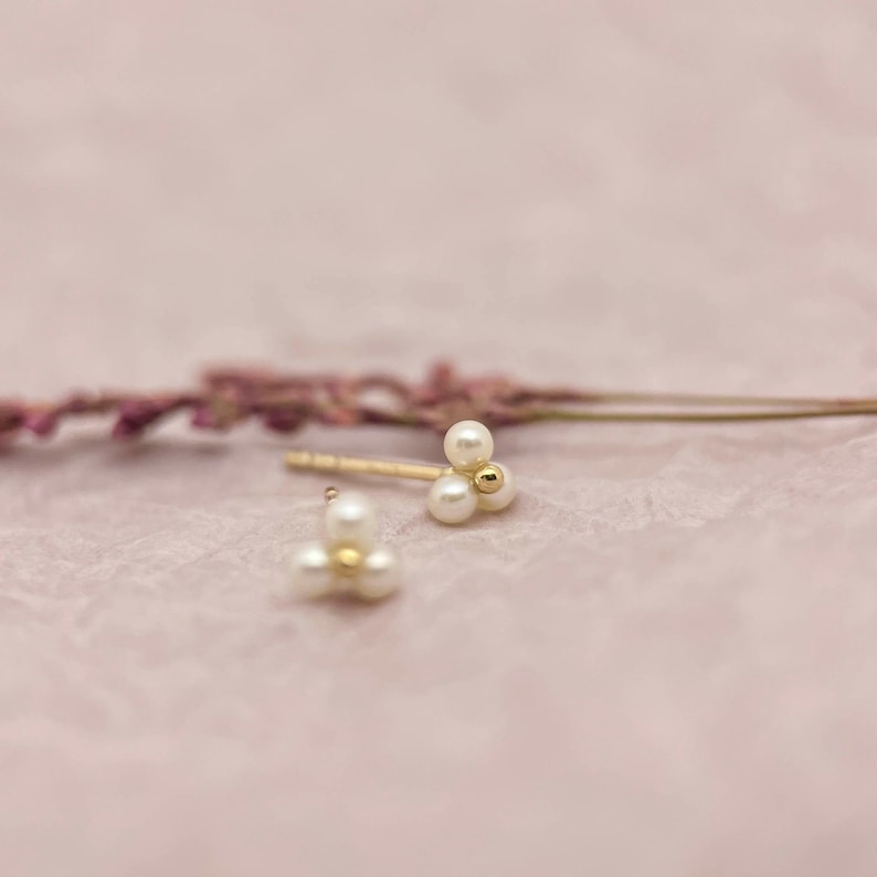 May include: Gold stud earrings, each with three small, white pearls and a central gold ball. The earrings are set against a light pink backdrop, with a blurred purple flower stem. The earrings are a delicate piece of jewellery.