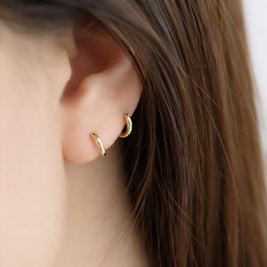 May include: Close-up of two small, gold hoop earrings on an ear. The earrings are simple, minimalist designs, with a polished, shiny finish. The earrings are worn in the earlobe and upper ear piercing. The background shows brown hair.