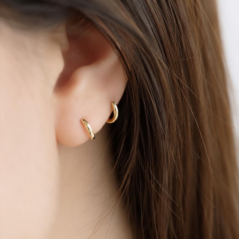 May include: Close-up of two small, gold hoop earrings on an ear. The earrings are simple, minimalist designs, with a polished, shiny finish. The earrings are worn in the earlobe and upper ear piercing. The background shows brown hair.