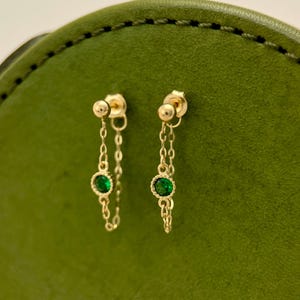 Op de afbeelding: Een paar gouden oorbellen met een delicate ketting en een kleine groene edelsteen. De oorbellen zijn op een groene achtergrond.