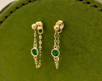 May include: A pair of gold earrings with a delicate chain and a small green gemstone. The earrings are on a green background.