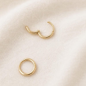 May include: Two gold-colored hoop earrings, one open and one closed, rest on a soft, off-white surface. The earrings have a simple, circular design, with a hinged clasp on the open earring. The jewelry is displayed in a well-lit setting.