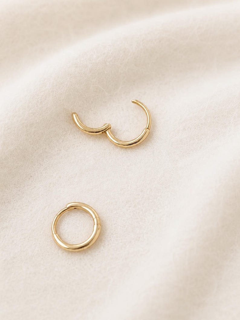 May include: Two gold-colored hoop earrings, one open and one closed, rest on a soft, off-white surface. The earrings have a simple, circular design, with a hinged clasp on the open earring. The jewelry is displayed in a well-lit setting.