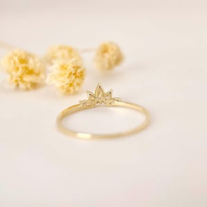 Puede incluir: Un delicado anillo de oro con dise&ntilde;o de corona. El anillo tiene una banda delgada y una parte superior detallada en forma de corona. El anillo se encuentra sobre un fondo suave y neutro con acentos florales amarillos borrosos, creando una est&eacute;tica minimalista.