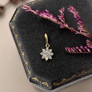 May include: A gold pendant featuring a central round clear gemstone surrounded by eight smaller round clear gemstones in a floral arrangement. The pendant includes a small gold bail at the top for attachment to a chain or necklace.