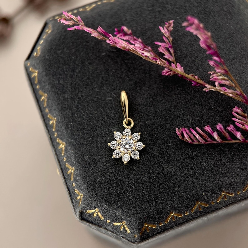 May include: A gold pendant featuring a central round clear gemstone surrounded by eight smaller round clear gemstones in a floral arrangement. The pendant includes a small gold bail at the top for attachment to a chain or necklace.