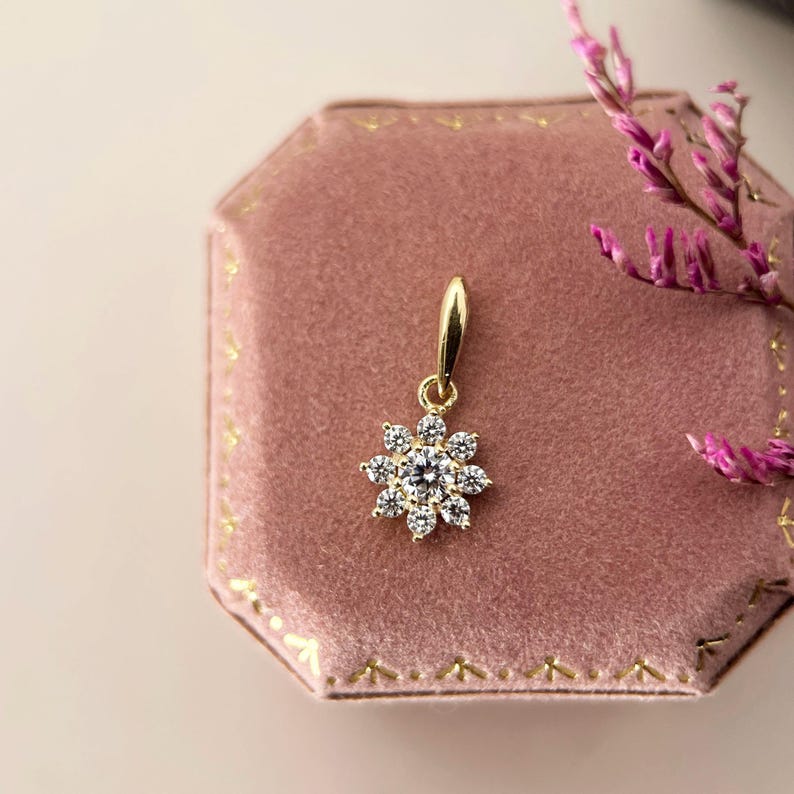 May include: A gold pendant featuring a central round clear gemstone surrounded by eight smaller clear gemstones arranged in a flower-like pattern. The pendant includes a gold bail at the top for attachment to a chain, showcasing a polished finish.