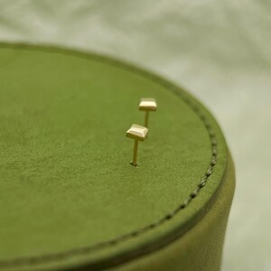 May include: Two gold square stud earrings on a green suede surface.
