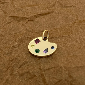 Op de afbeelding: Een gouden kunstenaars-paletbedel met gekleurde edelstenen. De bedel heeft een rode, gele, groene, blauwe en paarse edelsteen.