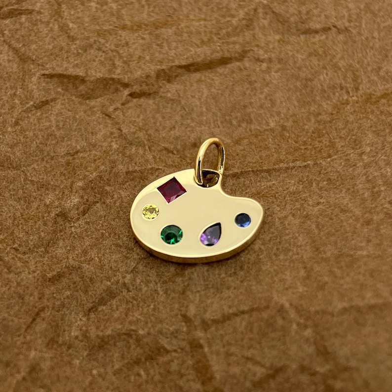 Op de afbeelding: Een gouden kunstenaars-paletbedel met gekleurde edelstenen. De bedel heeft een rode, gele, groene, blauwe en paarse edelsteen.