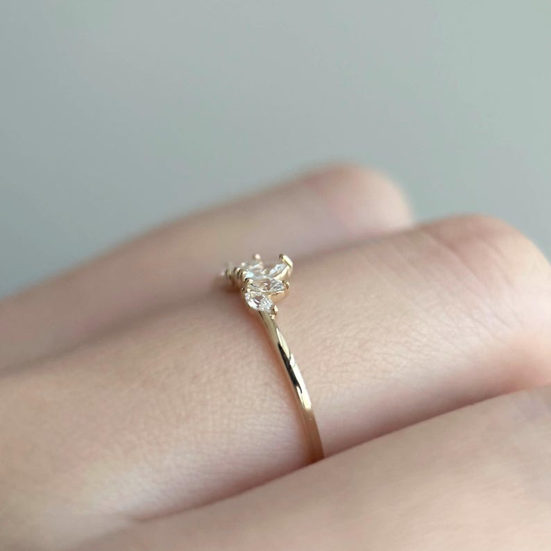 Puede incluir: Un delicado anillo de oro con un grupo de peque&ntilde;as piedras preciosas transparentes. La banda del anillo es fina y pulida, con las piedras preciosas dispuestas en un patr&oacute;n floral o de estrella. El anillo se muestra en un dedo.