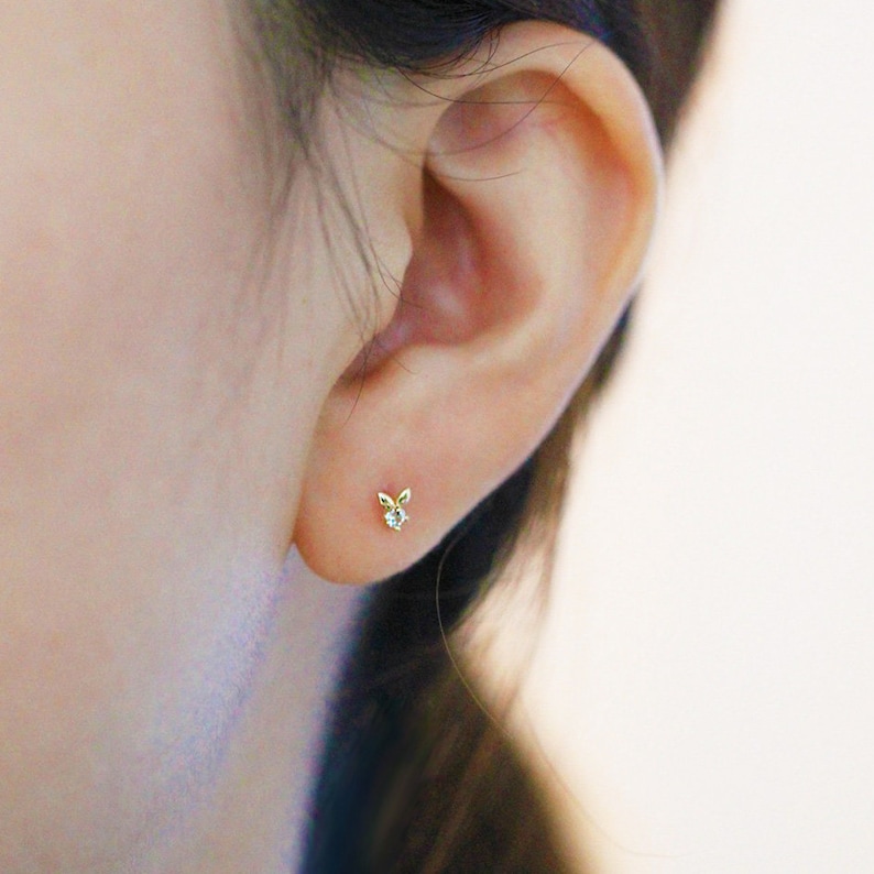 May include: A close-up of a person's ear with a small gold earring featuring a delicate bunny design with a single clear stone in the centre.