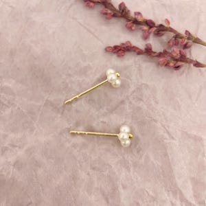 May include: A pair of gold-coloured stud earrings, each with a cluster of three small, white pearls. The earrings are set against a soft pink background, with a sprig of purple flowers in the upper right corner.