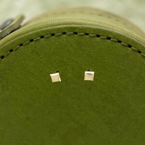 May include: Two small, square-shaped gold earrings on a green leather surface.