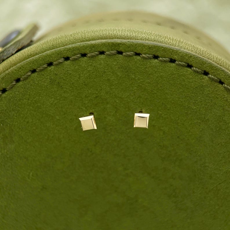May include: Two small, square-shaped gold earrings on a green leather surface.