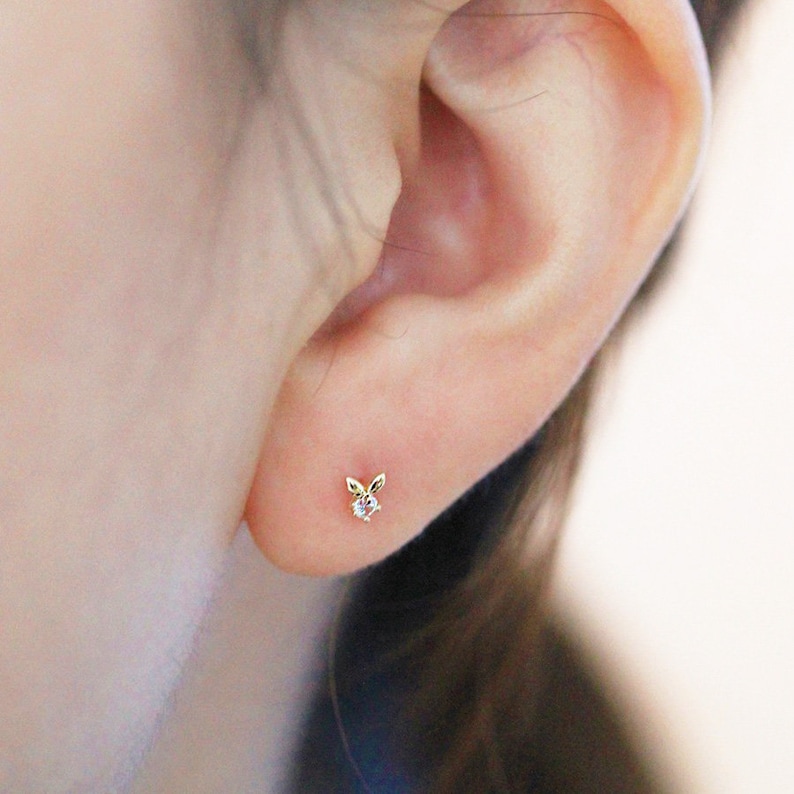 May include: A close-up of a person's ear with a small gold earring featuring a bunny rabbit design with a clear stone in the centre.