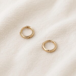 May include: Two small, gold-colored hoop earrings. The earrings are round with a hinged closure. The earrings are displayed on a white, textured surface. The earrings are a simple, classic design.