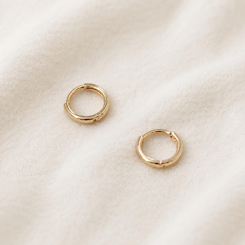 May include: Two small, gold-colored hoop earrings. The earrings are round with a hinged closure. The earrings are displayed on a white, textured surface. The earrings are a simple, classic design.