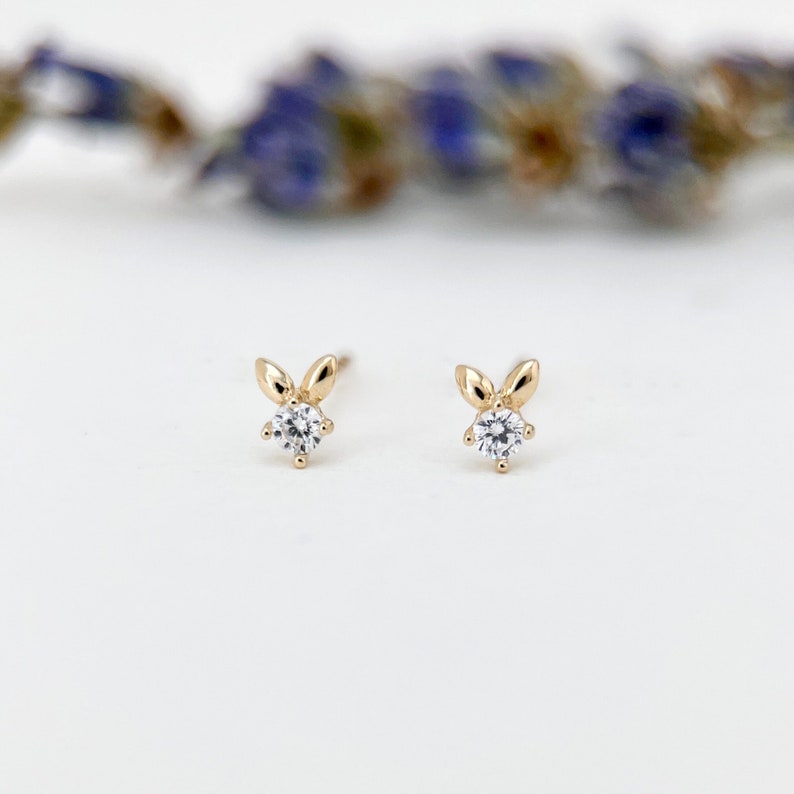 May include: A pair of gold stud earrings with a small, clear gemstone in the centre of each earring. The earrings are shaped like a bunny's ears.