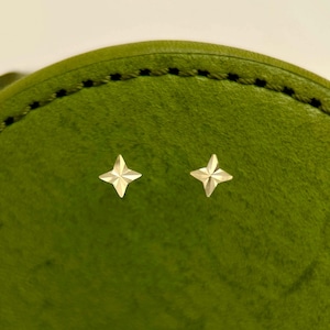 May include: A pair of small, gold-colored star-shaped stud earrings. The earrings are set against a textured, green background. The earrings are likely made of metal and are designed to be worn in pierced ears.
